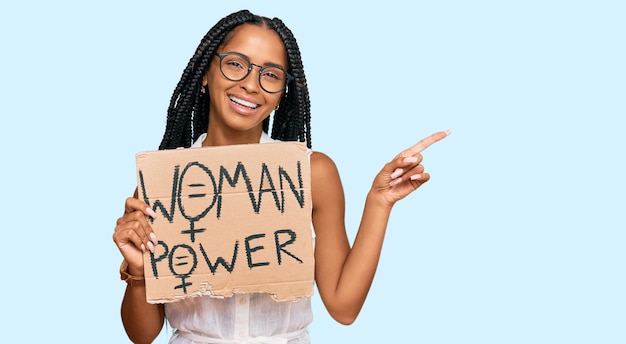 Beautiful hispanic woman holding woman power banner smiling happy pointing with hand and finger to the side