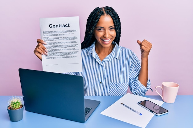 Beautiful hispanic woman at the office showing contract document screaming proud, celebrating victory and success very excited with raised arm