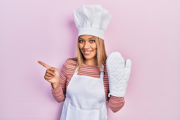 Beautiful hispanic woman uniform wearing apron and hat wearing protective glove smiling happy pointing with hand and finger to the side