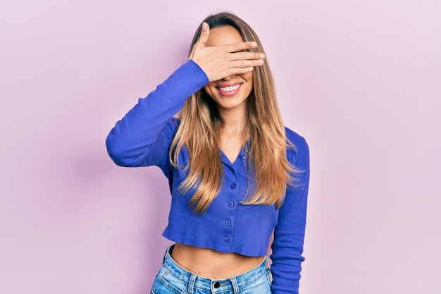 Beautiful hispanic woman wearing casual blue shirt smiling and laughing with hand on face covering eyes for surprise blind concept