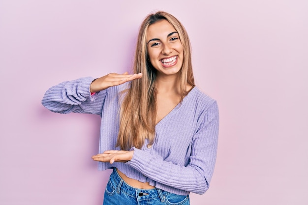 Foto bella donna ispanica che indossa una camicia casual gesticolando con le mani che mostrano il simbolo di misura del segno di grandi e grandi dimensioni sorridendo guardando il concetto di misurazione della fotocamera