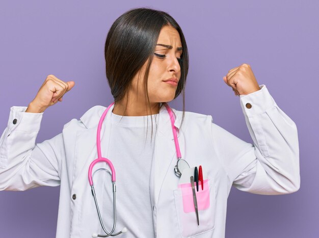 Beautiful hispanic woman wearing doctor uniform and stethoscope showing arms muscles smiling proud fitness concept