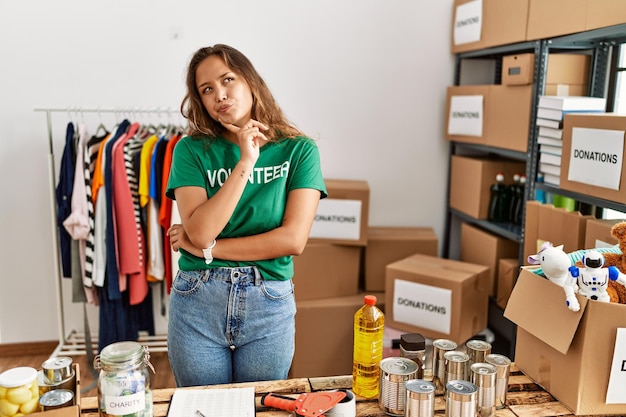 寄付スタンドでボランティアTシャツを着た美しいヒスパニック系女性は、あごに指を当てて疑いについて集中して考え、不思議に思いながら見上げている