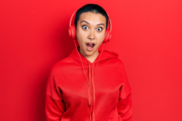 Beautiful hispanic woman with short hair listening to music using headphones afraid and shocked with surprise expression, fear and excited face.