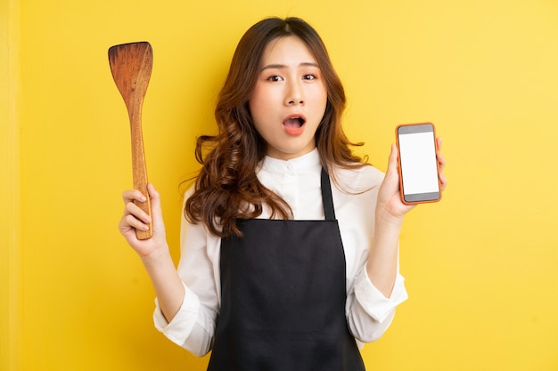 Beautiful housewife holding rice spoon and phone with happy surprise isolated on yellow