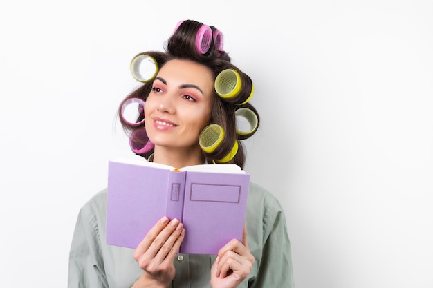 Beautiful housewife Young cheerful woman with curlers bright makeup with a book in her hands on a white background Thinking about a dinner recipe Looking for food ideas