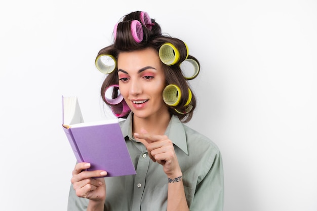 Bella casalinga giovane donna allegra con bigodini trucco luminoso con un libro in mano su uno sfondo bianco pensando a una ricetta per la cena alla ricerca di idee alimentari