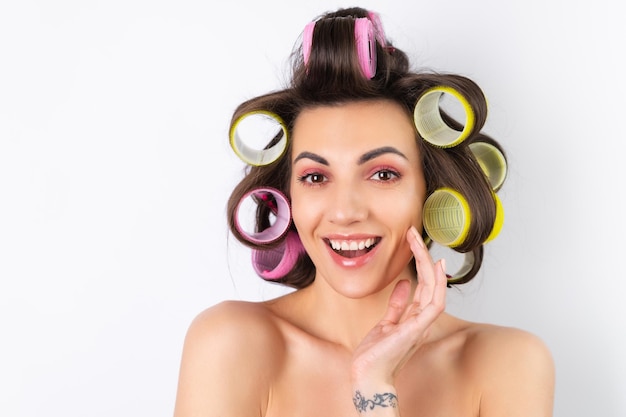 Beautiful housewife Young cheerful woman with hair curlers and bare shoulders getting ready for a date night Makes a hairstyle at home on a white background