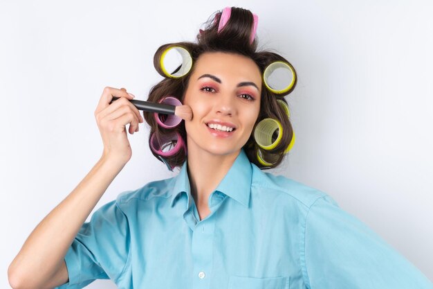 Beautiful housewife Young cheerful woman with hair curlers bright pink makeup and a cotouring brush is preparing for a date night dinner on a white background