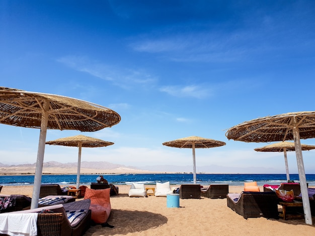 Foto bella immagine della spiaggia con lettini, lettini e pouf sotto gli ombrelloni di paglia in spiaggia contro il cielo blu