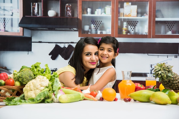 아름다운 인도 또는 아시아 젊은 엄마와 딸이 부엌에 있고 과일과 야채가 가득한 식탁이 있습니다.