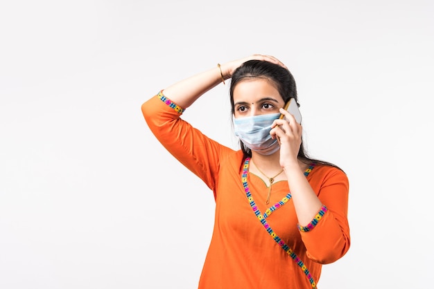 흰색 벽 위에 격리된 채 보호 의료 마스크를 착용한 채 휴대전화로 통화하는 아름다운 인도 여성