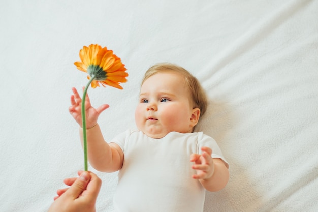 Beautiful infant baby lying on white sheets reaching for a flower.