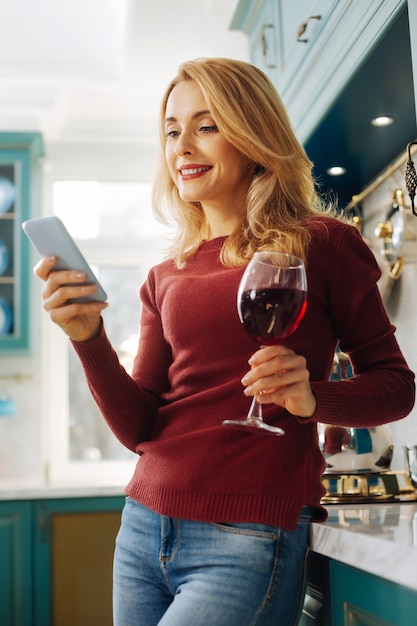 Beautiful inspired blond young woman smiling and holding a phone while drinking some red wine and relaxing