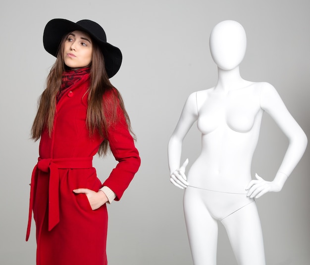 Beautiful interesting girl in the hat next to the white female mannequin on grey background
