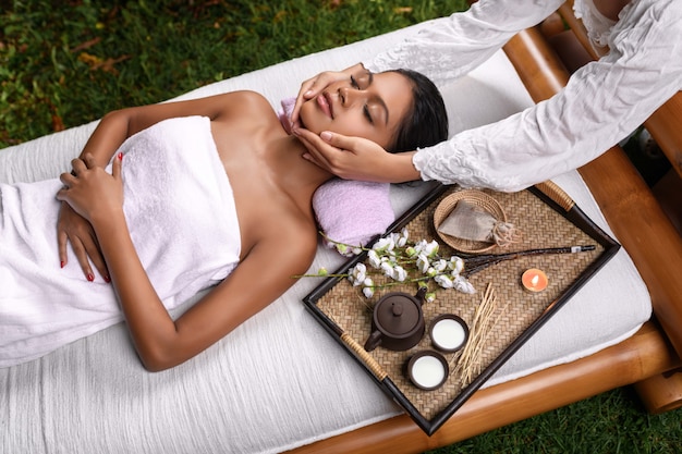 A beautiful interracial girl lies on a massage table on which stands a tray with aroma complex and receives a facial massage