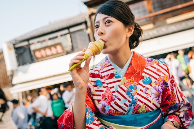 일본 거리에 서서 맛있는 말 아이스크림을 먹고 있는 아름다운 일본 여성. 디저트 컨셉을 먹는 꽃 기모노를 입은 일본 소녀. 일본의 여름 라이프 스타일.