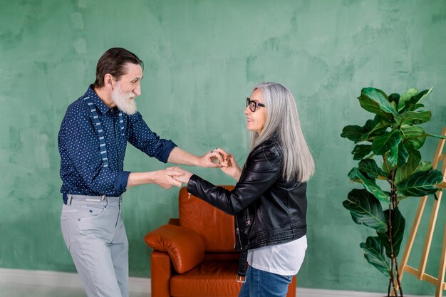 Beautiful joyful stylish senior woman with long straight gray hair, dancing together with her handsome bearded husband