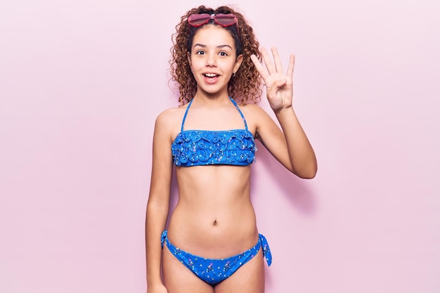 Beautiful kid girl with curly hair wearing bikini and sunglasses showing and pointing up with fingers number four while smiling confident and happy.