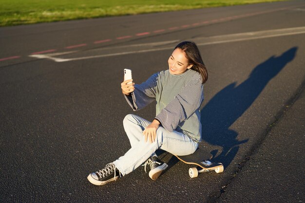 写真 美しい韓国人少女がスマートフォンでセルフィーを撮る スケートボードで写真を撮る 晴れた日を楽しむ人