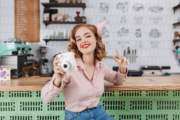 Beautiful lady in birthday cap sitting with white little camera in hand and happily showing victory gesture while spending time in cafe