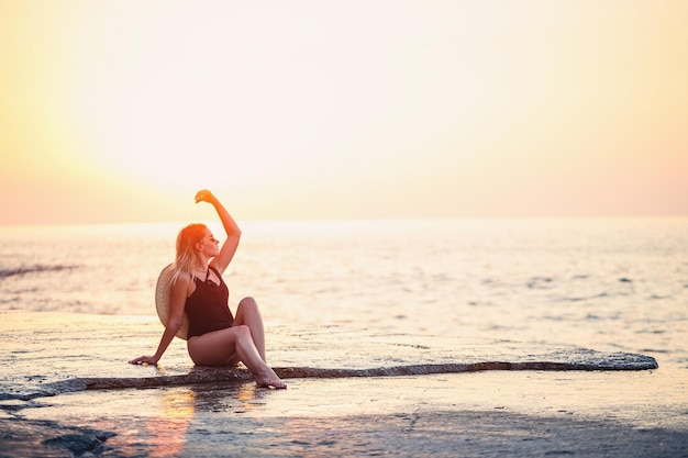 Beautiful lady in a black swimsuit on the seashore Smiling face with a straw hat on her shoulders Golden sunset light