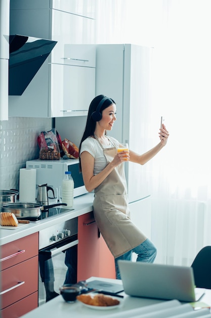 Foto bella signora in abiti casual e un grembiule in piedi con un bicchiere di succo d'arancia e sorridente mentre scatta foto