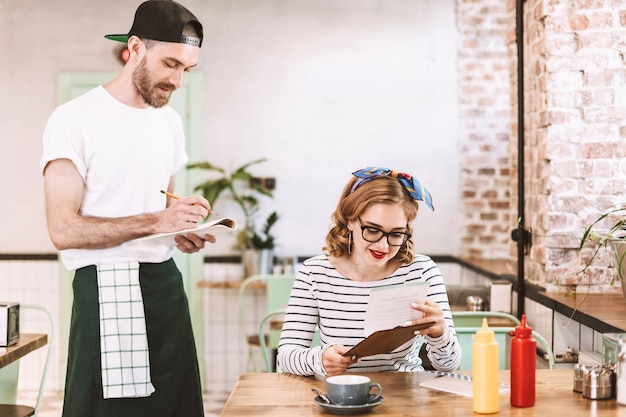 Beautiful lady in eyeglasses sitting at the table with menu in hands and making order while waiter standing near and writing in cafe