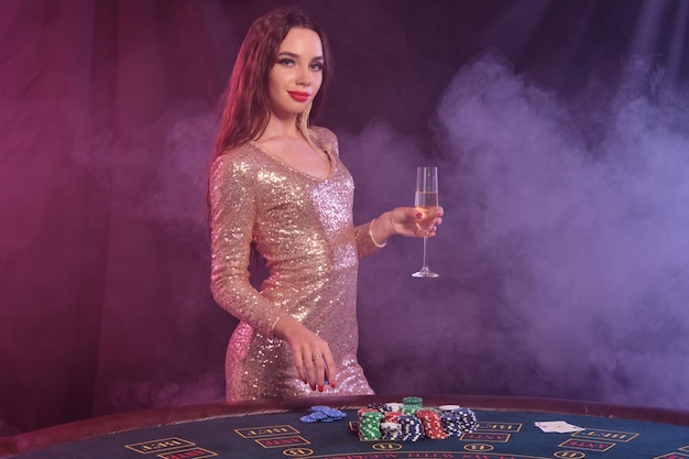 Beautiful lady in golden dress holding chip and glass of champagne posing sideways at playing table in casino Black smoke background colorful backlights Gambling entertainment poker Closeup