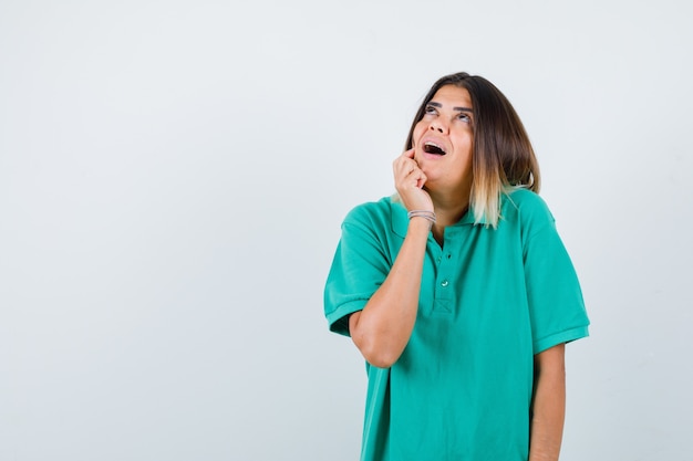 Beautiful lady holding finger on cheek, looking up in green polo t-shirt and looking wondered. front view.