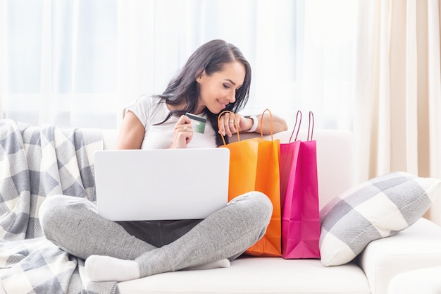 Beautiful lady looking into colorful paper shopping bags next to her smiling, with a laptop on her criss crossed legs and a payment card in her hand in a light living room.