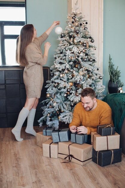 プレゼントを包む男を気遣いながらクリスマスツリーに飾りを置く美しい女性