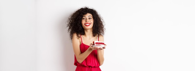 Красивая дама в красном платье празднует день рождения, держа кусок торта со свечой и улыбающуюся подставку