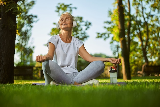 Bella signora in abbigliamento sportivo seduta su un tappetino da yoga e meditando mentre si gode le canzoni preferite all'aperto