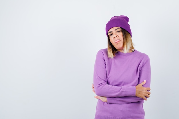 Foto bella signora in maglione, berretto con le mani incrociate, che tira fuori la lingua e sembra sicura, vista frontale.