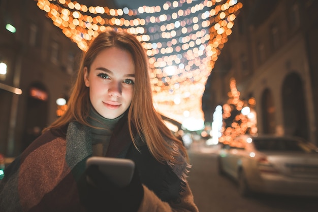 Beautiful lady in warm clothes stands with a smartphone in her hands at the background of a night street