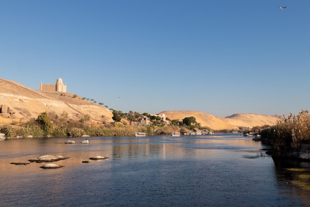 Beautiful landmark of Nile river bank shore with traditional construction village in the desert dunes and mountains with faluca boat sailing