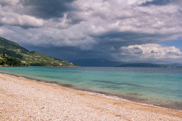 Красивый пейзаж - бирюзовая морская вода, золотой песок, серое небо с темными грозовыми облаками и горы на горизонте.