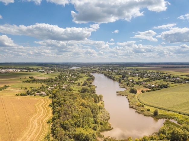 Beautiful landscape with agricultural fields, country and wide river on a background of blue cloudy sky in a summer sunny day. Aerial view from drone.