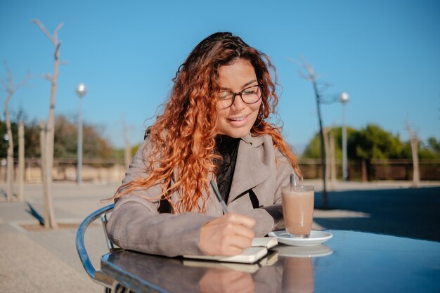 Bella ragazza latina che beve una tazza di caffè e guarda il telefono cellulare, su un tavolo di metallo all'aperto