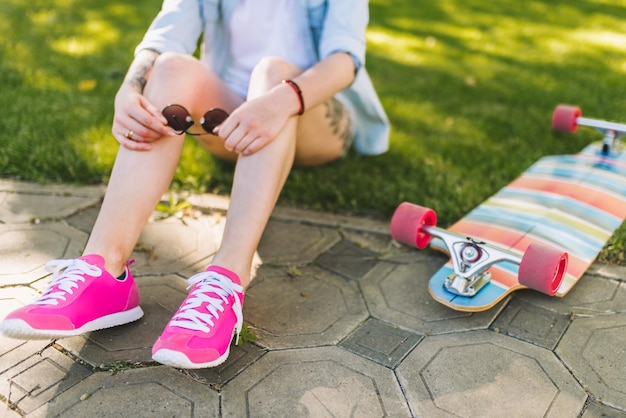 Photo beautiful legs of woman wearing pink shoes  with sunglasses in hand sitting in park on green grass background in a blue shirt sporty female relaxing after longboarding outdoor