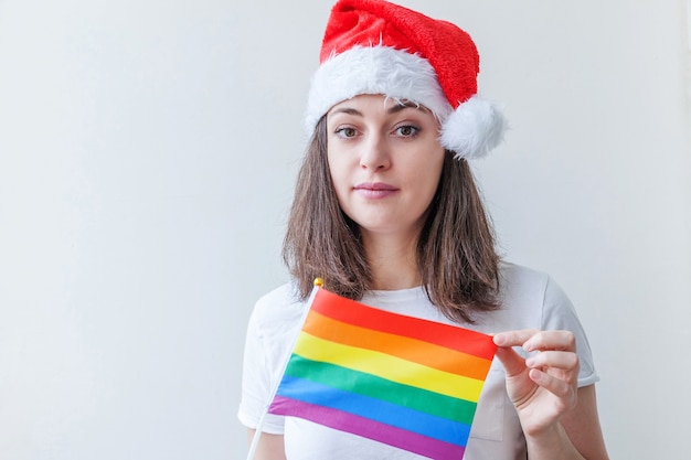 幸せで興奮しているように見える白い背景で隔離のLGBTの虹色の旗と赤いサンタクロースの帽子の美しいレズビアンの女の子。若い女性のゲイプライドの肖像画。ハッピークリスマスと年末年始。