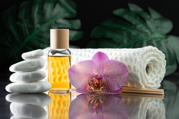 Bellissimo fiore di orchidea lilla, bottiglia trasparente di olio o profumo giallo, bastoncini di legno e asciugamano arrotolato con una pila di pietre bianche e foglie di monstera