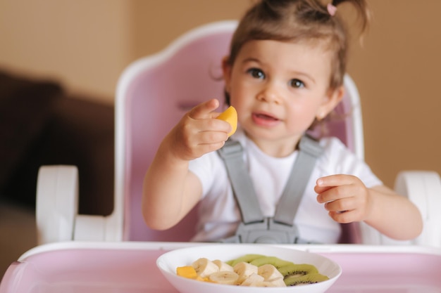 Photo beautiful little baby girl first time eating exotic fruits in high chair cute baby girl taste delitious fruist mango kiwi and babana