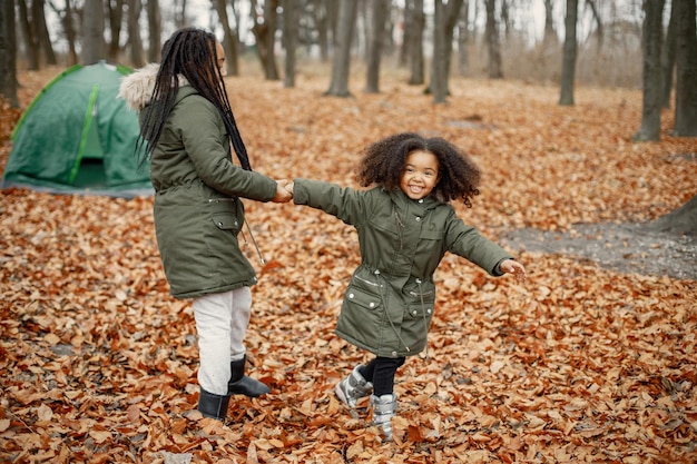 Beautiful little black girls standing near tent in the forest Two little sisters playing in autumn forest Black girls wearing khaki coats