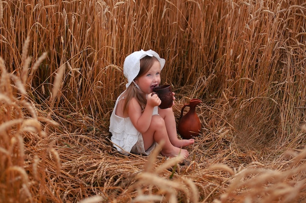 美しい小さなブロンドの女の子は、パンを食べて、ミルクを飲む麦畑に座っています