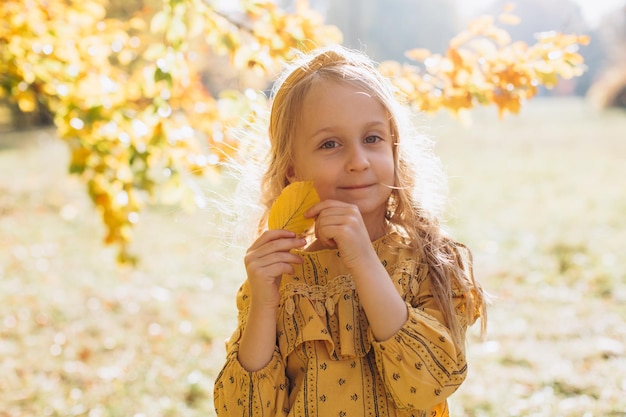 A beautiful little blonde girl is walking in an autumn park holding yellow maple leaves
