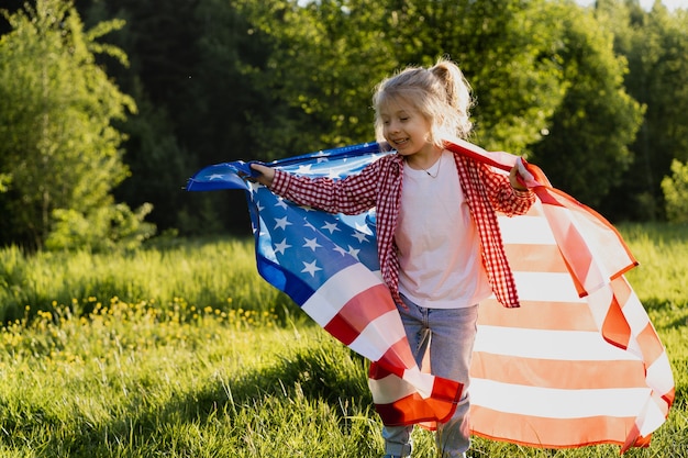 Beautiful little blonde girl with the American flag in nature in the sunlight.