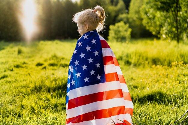 Beautiful little blonde girl with the American flag in nature in the sunlight.