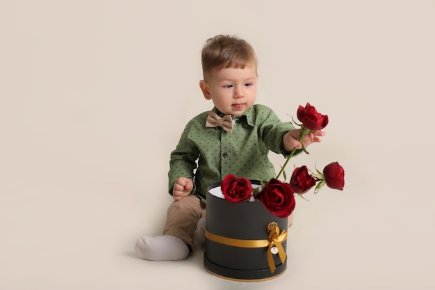 a beautiful little boy in a green shirt sits on a white background next to a box with red roses